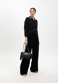 Schwarzer Strickpullover mit einem Nietenkragen, kombiniert mit schwarzen Samt-Hosen im weit geschnittenen Stil. Hält eine schwarze Handtasche aus Kunstfell mit einem goldenen "M".