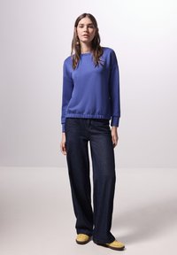 Blauwe sweatshirt met ribbelboord en manchetten, gecombineerd met donkere wijde jeans. Model draagt gele schoenen en staat tegen een neutrale achtergrond.