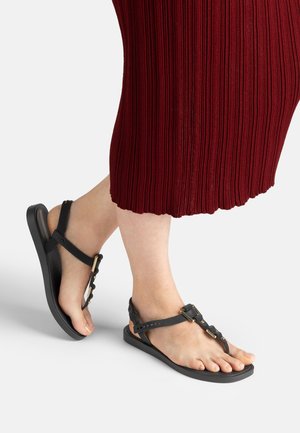 Füße, die schwarze flache Sandalen mit goldenen Schnallen tragen, kombiniert mit einem gerippten bordeauxroten Rock, der unterhalb der Knie endet.