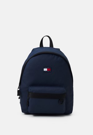 Sac à dos bleu marine avec poche zippée à l'avant, poignée noire et patch logo Tommy Jeans sur fond blanc uni.