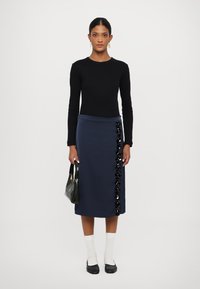 Tums zila midi svārki ar teksturētām melnām rotājumiem sānos, apvienoti ar pieguļošu melnu garām piedurknēm blūzi un melnu rokassomu.