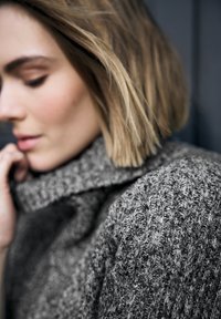 Grauer Strickpullover mit hohem Kragen, der ein strukturiertes Muster und dicken, kuscheligen Stoff aufweist. Das Modell berührt den Kragen mit der Hand.