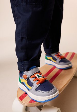 Piedi di bambino che indossano sneaker colorate con ciondoli a forma di dinosauro, in piedi su uno skateboard di legno con strisce rosse su uno sfondo semplice.