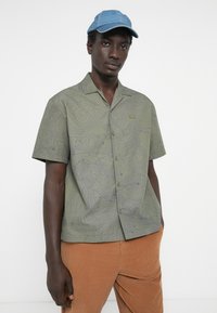 Short-sleeve green shirt with purple topographic pattern, collared design, front buttons, and an embroidered logo, paired with corduroy pants.