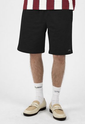 Person trägt schwarze Shorts, weiße Crew-Socken mit Text, beige geflochtene Loafer und ein rot-weiß gestreiftes Hemd, das an der Taille sichtbar ist.