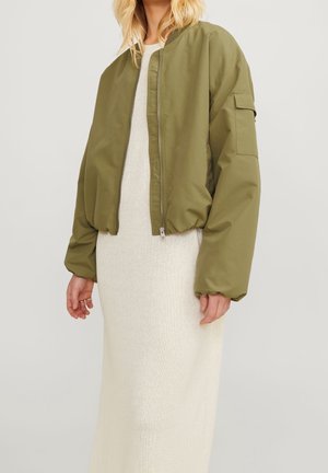 Frau trägt eine olivgrüne Bomberjacke über einem langen, cremefarbenen Strickkleid und steht vor einem schlichten, hellen Hintergrund.