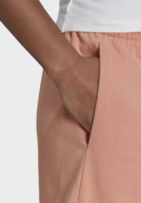 Růžové bavlněné tepláky se bočními kapsami, které mají měkkou texturu a elastický pás. Detailní záběr ukazuje ruku odpočívající v kapse.