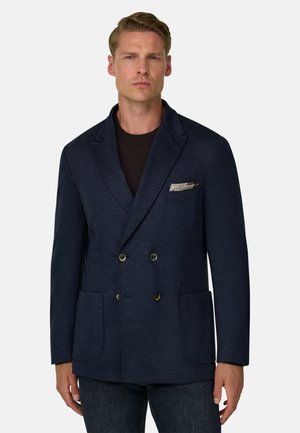 Homme portant un blazer croisé bleu marine avec un mouchoir de poche à motif et un jean foncé sur un fond uni.