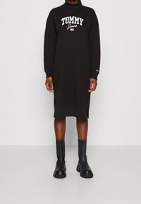 Czarna, oversize'owa sukienka-sweterek z wysokim kołnierzem, z białym logo "TOMMY Jeans". Posiada długie rękawy i boczne rozcięcia, zestawiona z czarnymi botkami.