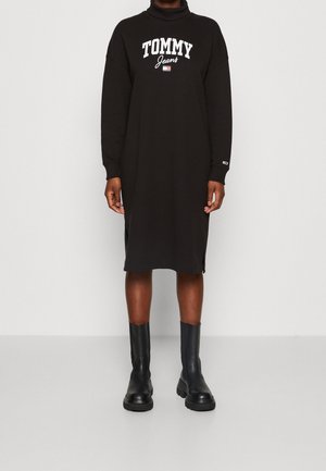 Robe-sweat à capuche noire oversize avec un col haut, ornée du logo blanc "TOMMY Jeans". Elle possède des manches longues et des fentes sur les côtés, associée à des bottes noires.