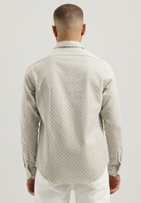 Man met kort haar, gekleed in een lichtbeige gestreept overhemd met lange mouwen en witte broek, die met zijn rug naar de camera staat tegen een effen achtergrond.