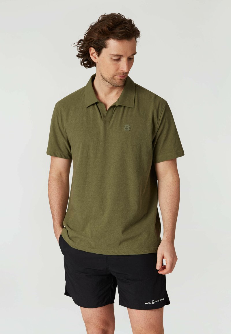 Olivgrön polo-shirt med en lös passform, korta ärmar och spetsig krage. Har en diskret logotyp på bröstet. Kombinerad med svarta shorts.