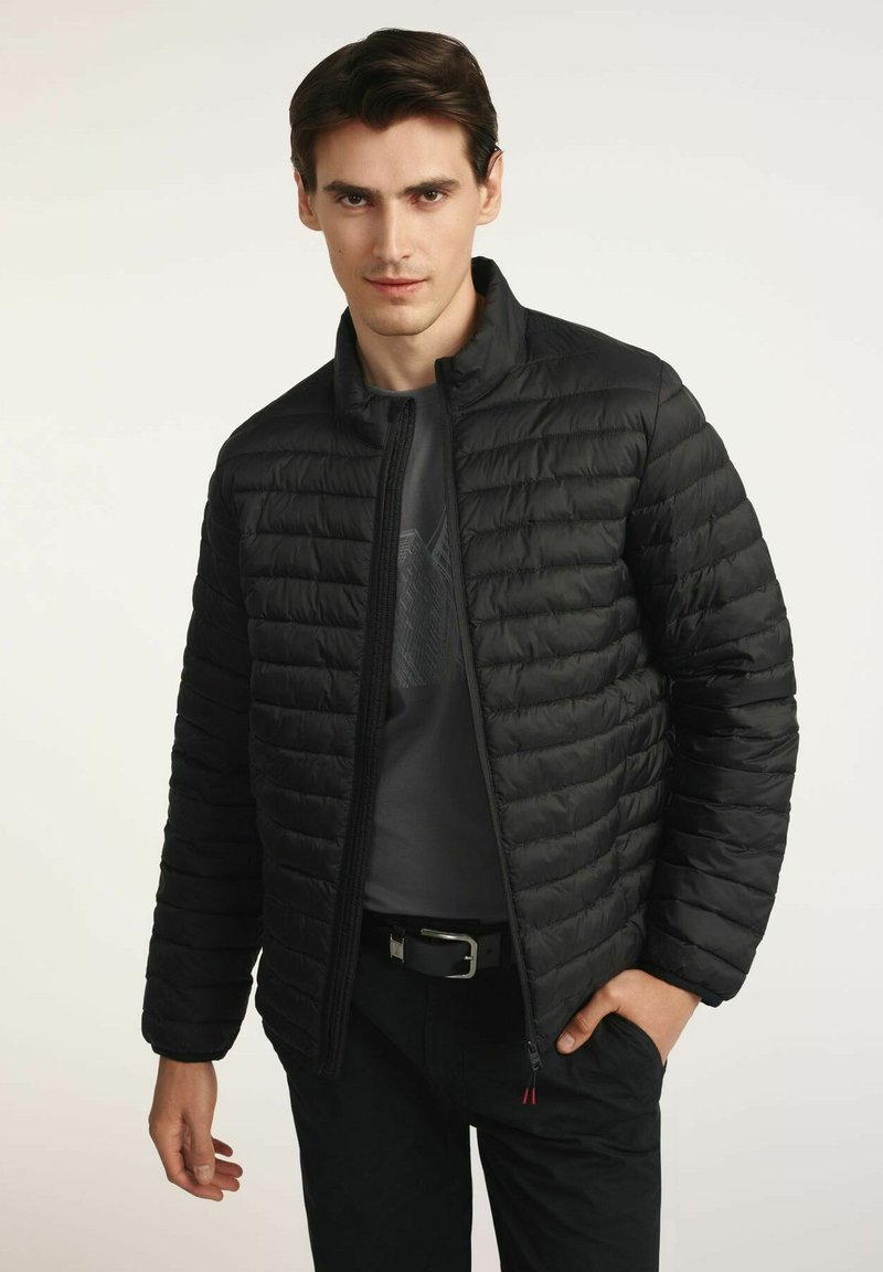 Czarna kurtka puchowa z wysokim kołnierzem, pikowanym wzorem, zapięciem na zamek z przodu oraz dwiema kieszeniami. Noszona na ciemną koszulę z graficznym wzorem.