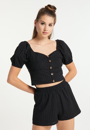 Giovane donna con capelli raccolti in uno chignon indossa un crop top nero con bottoni e maniche a sbuffo e pantaloncini neri elasticizzati in vita, posando con le mani sui fianchi.