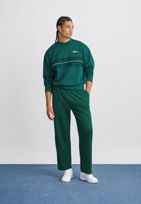 Zaļš vilnas džemperis ar baltām strīpām, kombinēts ar atbilstošām zaļām sporta biksēm. Abiem apģērba gabaliem ir brīvs griezums un elastīgs jostasvieta.