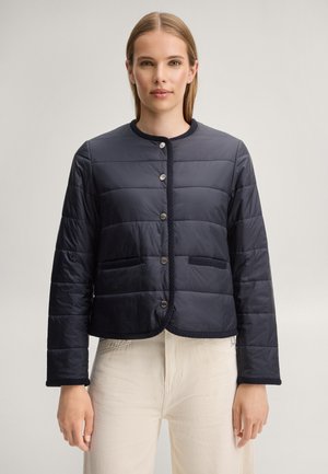 Femme portant une veste matelassée noire boutonnée avec un col rond et un pantalon crème taille haute, debout devant un fond uni.