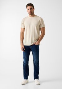 Uomo in piedi che indossa una maglietta beige a maniche corte, jeans blu scuro e scarpe da ginnastica bianche contro uno sfondo chiaro e semplice.