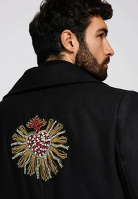 Schwarzer Mantel mit einem kunstvollen bestickten Design auf der Rückseite, das ein Herz zeigt, umgeben von goldenen Strahlen und roten Akzenten.