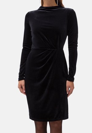 Długa sukienka z czarnego aksamitu z długim rękawem, sięgająca do kolan, z delikatnym drapowaniem w talii, noszona przez stojącą osobę z rękami swobodnie opuszczonymi wzdłuż ciała.