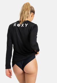 Long-sleeve black rash guard with white "ROXY" logo on the back, worn with black textured swim bottoms. Smooth fabric, casual fit.