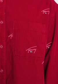 Red corduroy shirt featuring a button-up front, breast pocket, and embroidered logo. Texture is ribbed; buttons are round with a slight sheen.