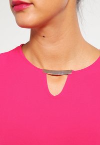 Top rosa brillante con scollatura a V e un accento metallico sul colletto. Tessuto liscio con maniche corte e vestibilità comoda.