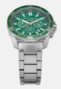 Ura zeleni cadran s srebrnim zapestjem, ki ima teksturirano površino, poglečne označbe ur, pod-števce in indikator vodoodpornosti.