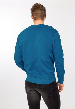 Turkos crewneck-tröja med långa ärmar och ribbad fåll, kombinerad med mörka slim-fit-jeans. Slät tyg utan synliga mönster.