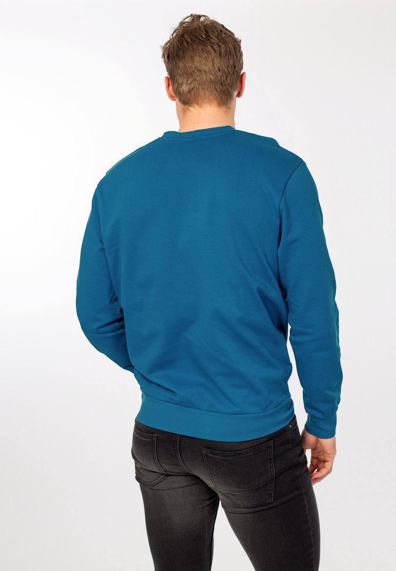 Sweat-shirt col ras du cou bleu sarcelle à manches longues avec un ourlet côtelé, associé à un jean slim foncé. Tissu lisse sans motifs visibles.