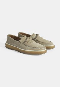 Zijden instapschoenen in lichtbeige met een rubberen zool, voorzien van decoratieve franjes en witte stikdetails langs de rand.