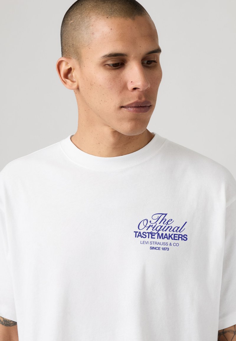 Giovane uomo con capelli corti che indossa una camicia bianca con testo blu "The Original Taste Makers Levi Strauss & Co Since 1873" sul petto.