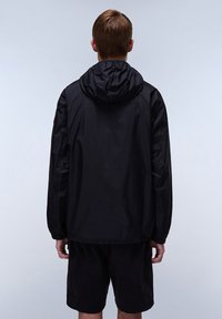 Zwarte lichtgewicht jas met capuchon, voorzien van elastische manchetten, een iets losse pasvorm en een gladde textuur. Draagt over donkere shorts.