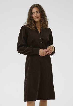 Femme aux cheveux bouclés, portant une robe marron foncé à manches longues avec des boutons, debout, les mains jointes devant un fond clair uni.