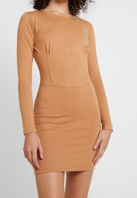 Długo- rękawowy sukienka typu bodycon w jasnobrązowym materiale, z dopasowaną górą i wyraźnie zarysowanymi szwami oraz gładką teksturą.