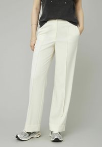 Pantalons blancs larges en tissu lisse, avec des plis avant et des ourlets retroussés, associés à des chaussures de sport argentées.