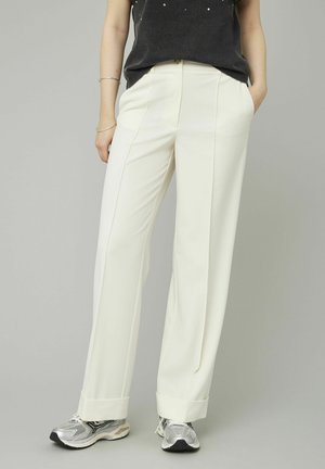 Pantalons blancs larges en tissu lisse, avec des plis avant et des ourlets retroussés, associés à des chaussures de sport argentées.