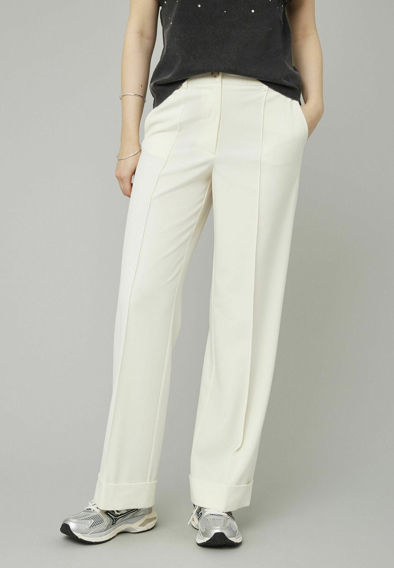Pantalons blancs larges en tissu lisse, avec des plis avant et des ourlets retroussés, associés à des chaussures de sport argentées.