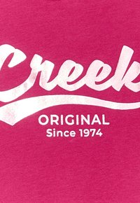 T-shirt di cotone rosa con testo bianco in grassetto: "Creek", con una sottolineatura curva, e "ORIGINAL Dal 1974" sotto di essa. Texture morbida.