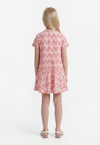 Ξανθό παιδί στέκεται με την πλάτη, φοράει κοντομάνικο φόρεμα με μοτίβο ζιγκ-ζαγκ σε ροζ, κόκκινο και λευκό και λευκά σανδάλια σε λευκό φόντο.