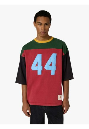 Giovane uomo che indossa una camicia a blocchi di colore, verde, rossa e nera, con un ampio numero 44 celeste chiaro sul davanti.