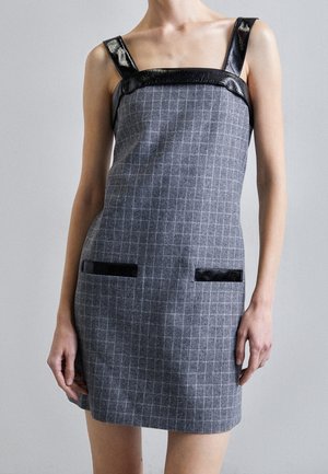 Szara sukienka bez rękawów w kratę, z czarnymi skórzanymi ramiączkami i kieszeniami, noszona przez osobę z rękami swobodnie opuszczonymi wzdłuż ciała.