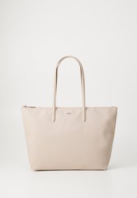 Sac tote beige en matériau texturé, avec deux anses allongées et une fermeture éclair. Logo Lacoste en accent sur le devant.