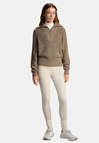 Brązowy sweter dzianinowy z półzamkiem, prążkowaną teksturą i luźnym krojem, w połączeniu z kremowymi legginsami i jasnymi sneakersami.