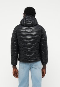 Zwarte pufferjack met capuchon, voorzien van een gewatteerd patroon en glanzende afwerking. Draagt men over lichtblauwe jeans.