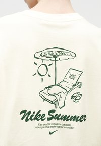 Πίσω μέρος κρεμ πουκάμισου με πράσινο γραφικό που απεικονίζει ήλιο, ομπρέλα παραλίας με την επιγραφή "La Dolce Vita", ξαπλώστρα με την ένδειξη "Nike Club", σανδάλια και το κείμενο "Nike Summer".