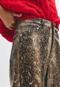 Pantaloni con motivo a pelle di serpente e finitura lucida, caratterizzati da un design aderente, dettagli in metallo argentato e vita alta, abbinati a un maglione rosso dalla texture. 