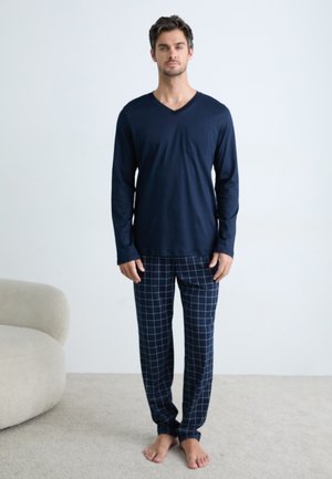Man staat blootsvoets in een marineblauw, langemouw V-hals shirt en marineblauwe geruite pyjamabroek op beige tapijt naast een gebogen bank.