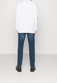 Top bianco a maniche lunghe con stampe colorate sulle maniche, abbinato a jeans di denim blu e scarpe nere, visto da dietro.