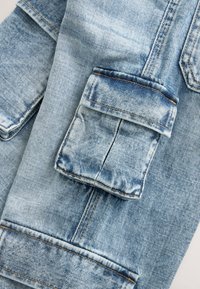 Manica di giacca di jeans azzurro chiaro con tasca a patta visibile e dettagli di cuciture arancione-marroni su tessuto strutturato.
