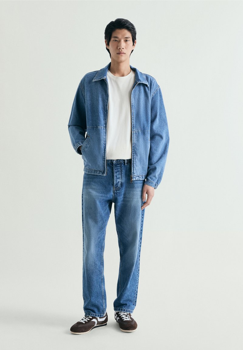 Ung mand iført blå denimjakke og jeans med hvid T-shirt og sorte sneakers mod en ensfarvet lys baggrund.